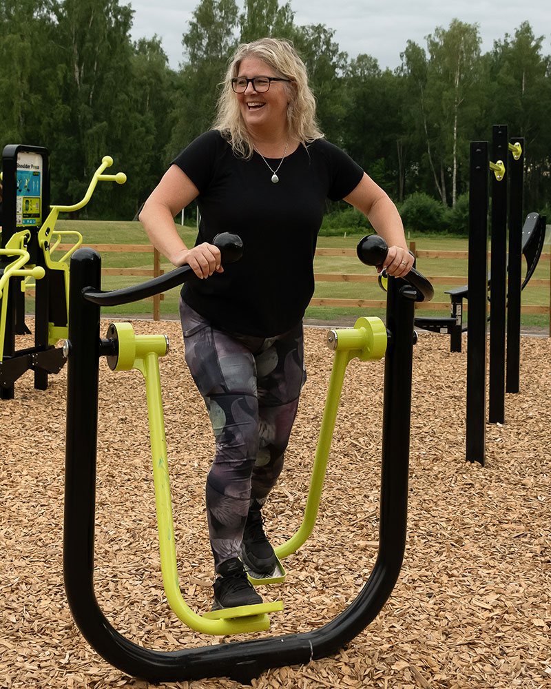 A woman exercises on an outdoor elliptical exercise machine, she is looking to the side and smiling.